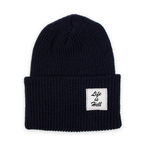 Photo of the "Life is Hell" Beanie by No Fun®.  Product is photographed against a white background.  Beanie is navy blue, and features a small white woven label on the cuff that reads "Life Is Hell".  Label is white, with black text, and red contrasting stitching.