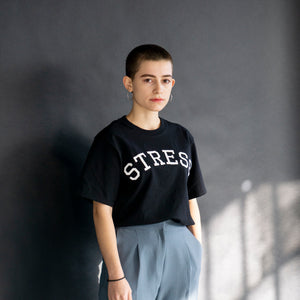 Photo of a model wearing the original NO FUN® "STRESS" t-shirt. Shirt is black and features large white collegiate style text that reads "STRESS" across the front in an arc shape.