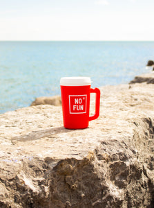 "Big Sipper" Mug in red by No Fun®.  The mug is sitting on top of a large stone, and a lake can be seen behind it. 