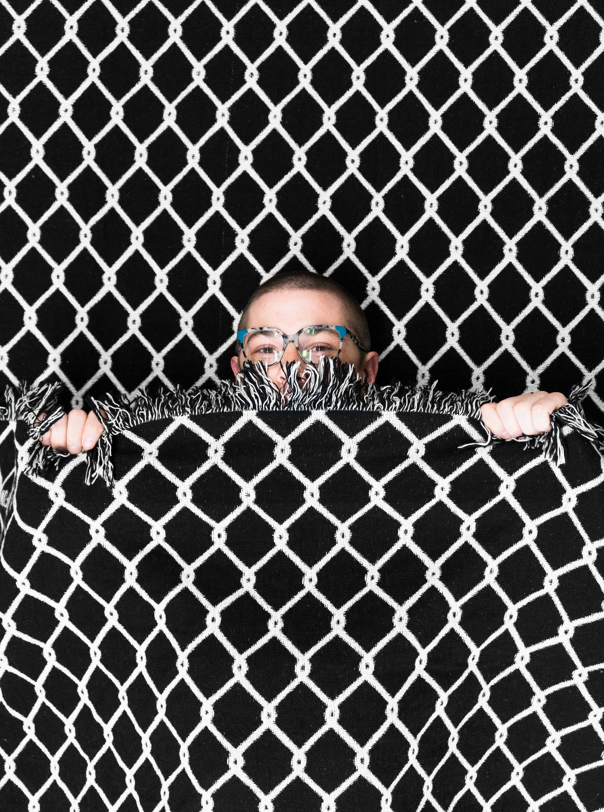 A person underneath the "Chainlink" woven blanket by No Fun®.  The person is peaking over the top of the blanket to showcase the scale of the design.  Only the top half of their face, and hands, are visible.