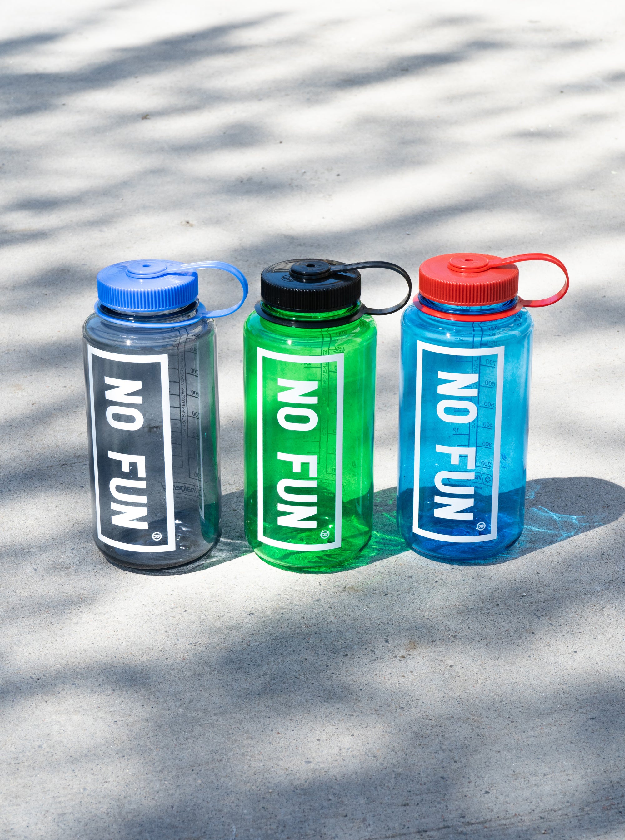3 colours of the Nalgene® + No Fun® 32oz Bottle.  They are lined up in a row on the concrete.  Shadows of leaves can be seen cast over the bottles.