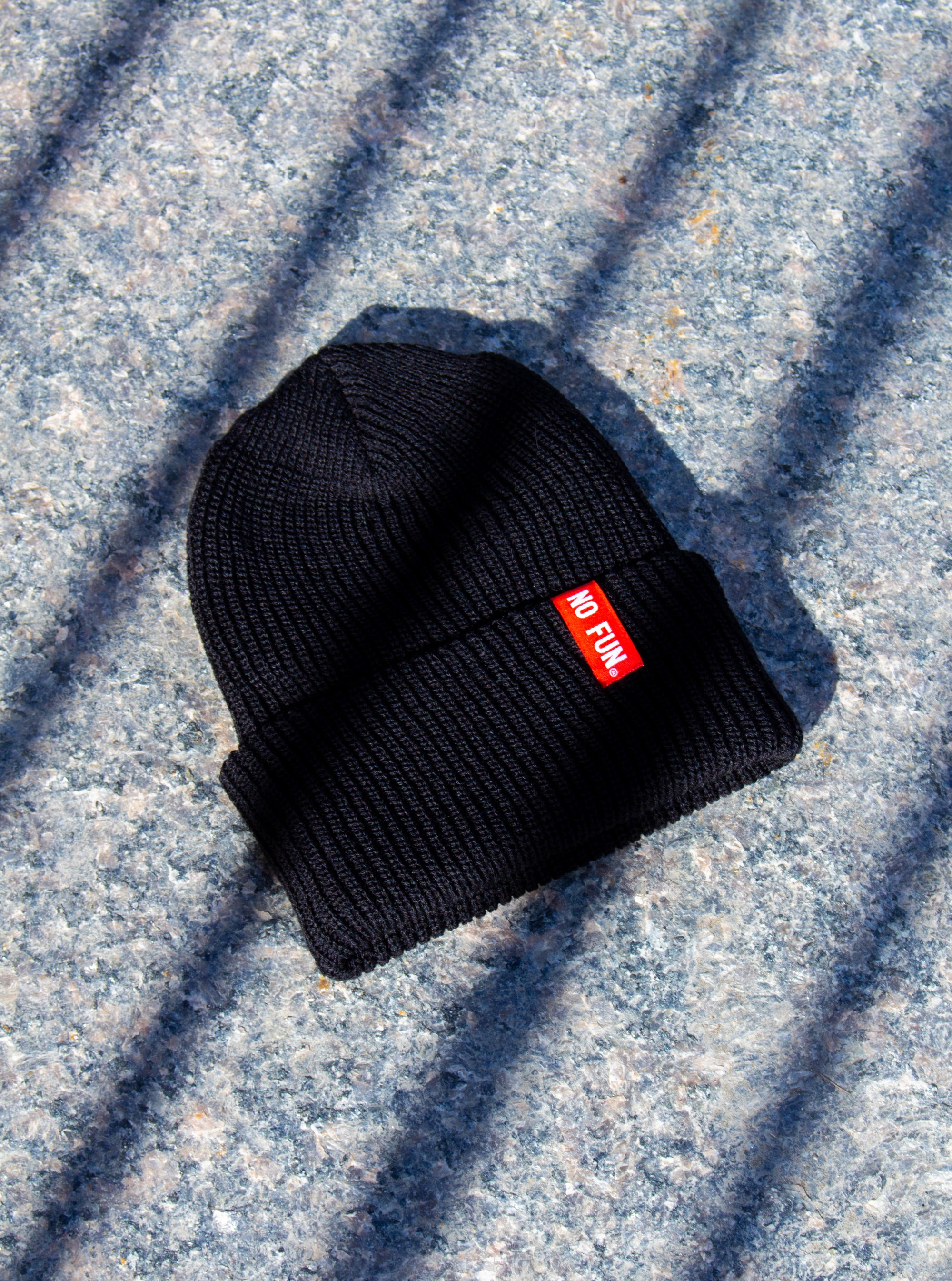 Photo of the "No Fun®" Ribbed beanie photographed on a slab of concrete, with diagonal shadows being cast upon the product.  Beanie is Navy Blue, and features a small, red, woven label that folds over the cuff. The text on the red label says "No Fun®" in white.