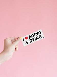 Hand holding the original, white, "Aging & Dying" bumper sticker.