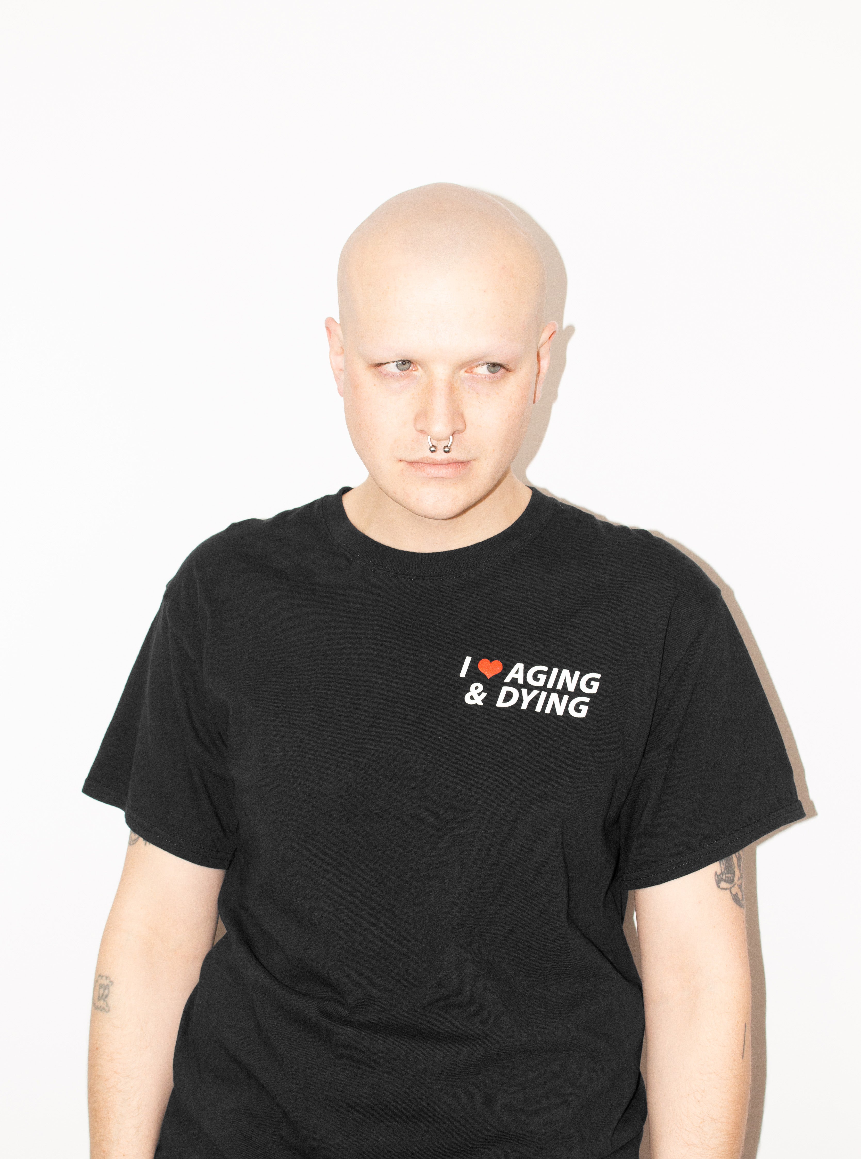 The original "Aging & Dying" T-shirt by No Fun®.  T-shirt is black, and is photographed on a model standing against a white background.  The left chest print text reads "I ❤️ Aging & Dying" and is located on the front of the T-shirt.   The text is white, with a red heart.