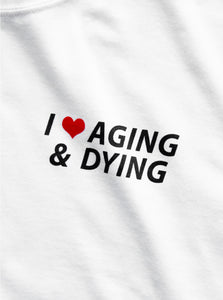 The original "Aging & Dying" T-shirt by No Fun®.  T-shirt is white, and is photographed close up to show the print detail.  The print text reads "I ❤️ Aging & Dying" and is located on the front of the T-shirt.   The text is black, with a red heart.