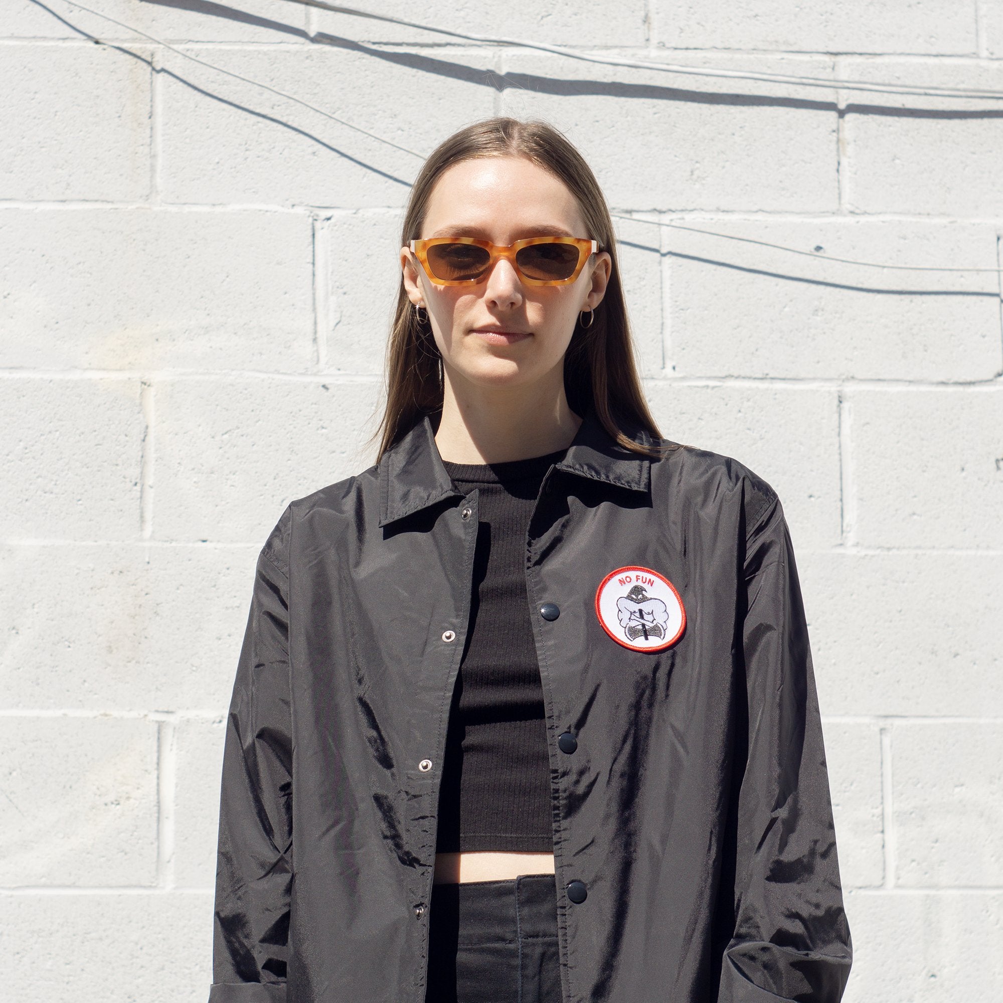 Photo of a female model wearing the "NO FUN®" X "Crawling Death" collaboration coaches jacket.  The model is showcasing front of the jacket with the black, red, and white executioner patch and snap closures.