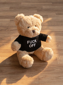 The "Friendly" Teddy Bear by No Fun®. Plush toy bear is beige and wears a black shirt with the phrase "FUCK OFF" printed on the front. Bear has a brown nose, and brown plastic eyes. Bear is photographed placed on a wood floor.