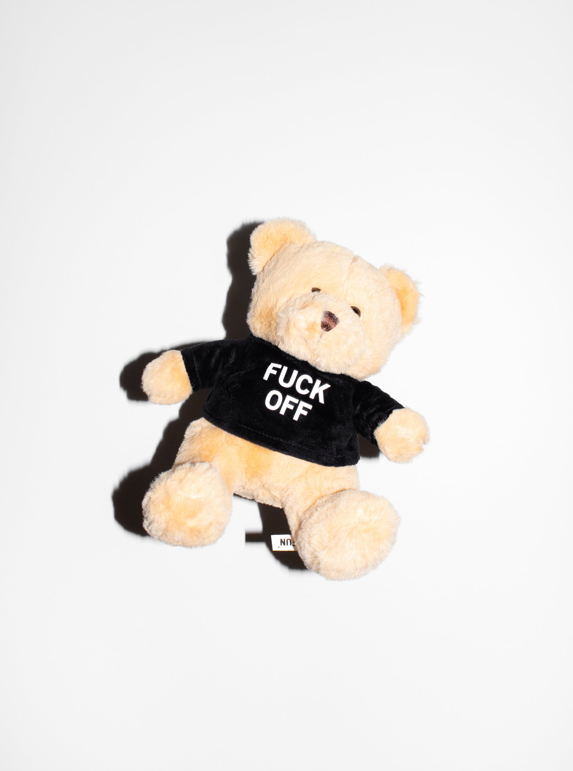 The "Friendly" Teddy Bear by No Fun®. Plush toy bear is beige and wears a black shirt with the phrase "FUCK OFF" printed on the front. Bear has a brown nose, and brown plastic eyes.  Bear is photographed floating against a white background.