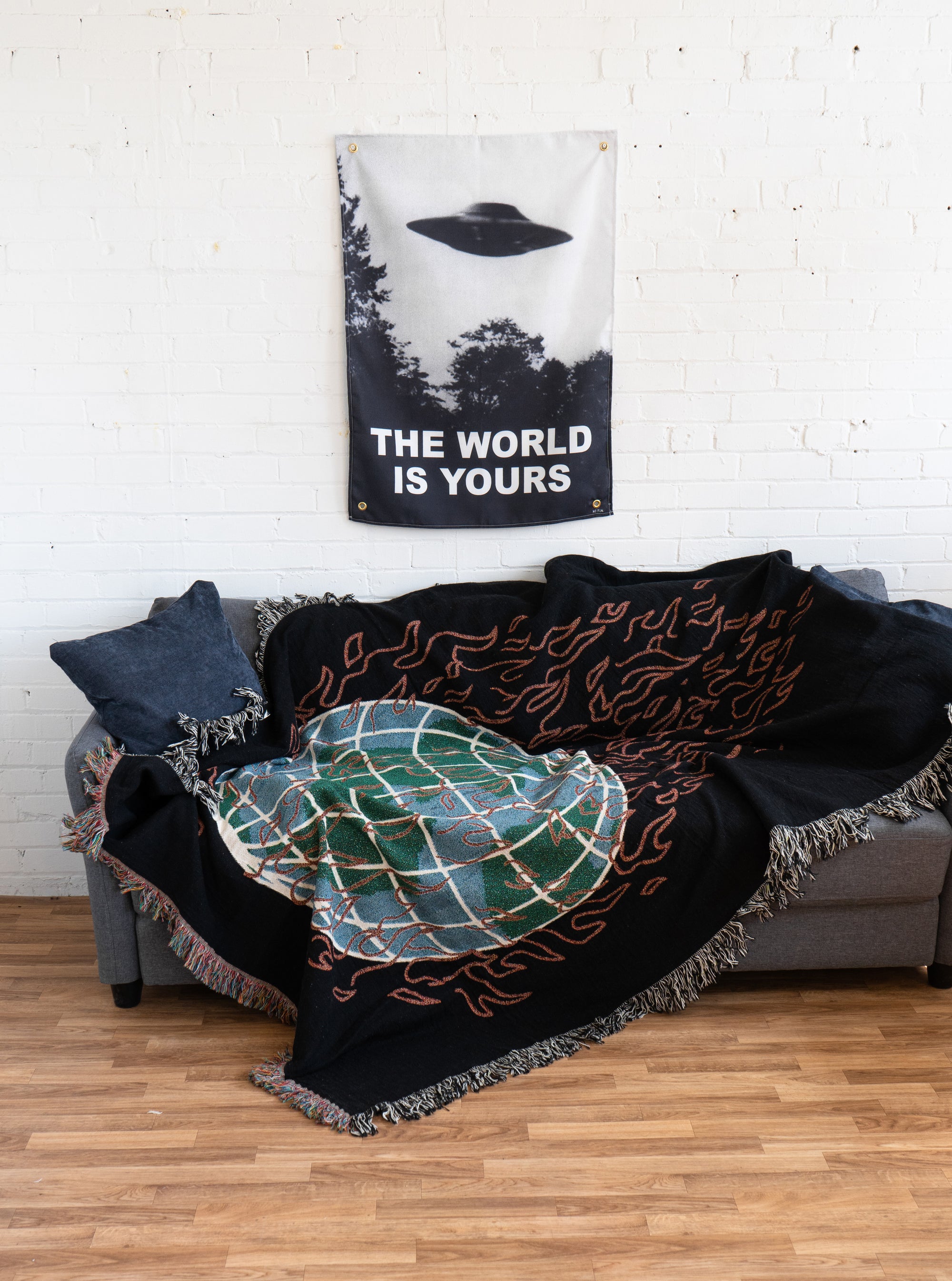 The "Global Warming" Woven Blanket by No Fun® on a grey couch in an untidy manner.  The couch is in a white room that has hardwood floor.  There is a wall tapestry hanging on the wall above the couch.