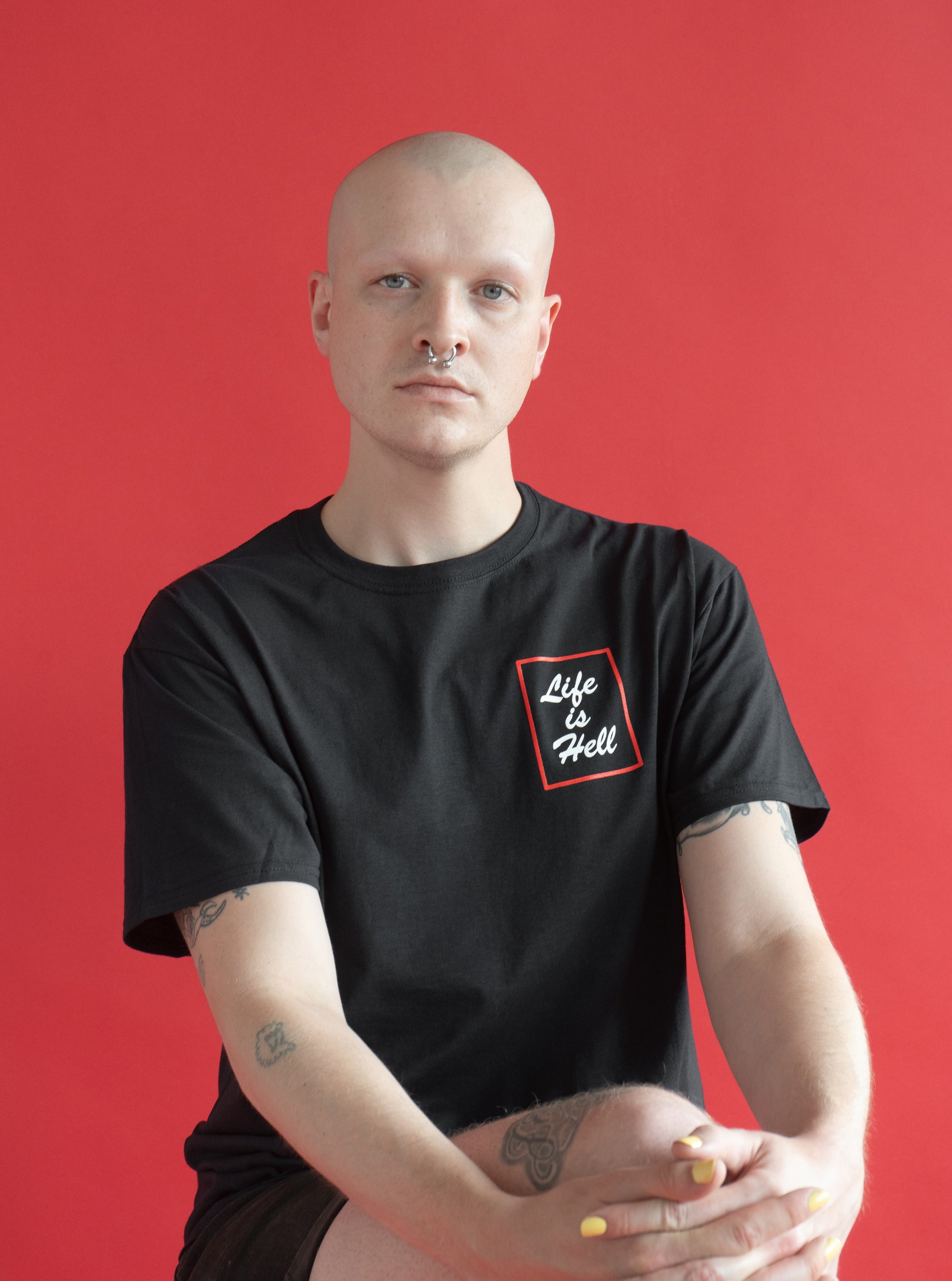 Model wearing the "Life is Hell" T shirt.  They have a shaved head, and are sitting in front of a red background.  