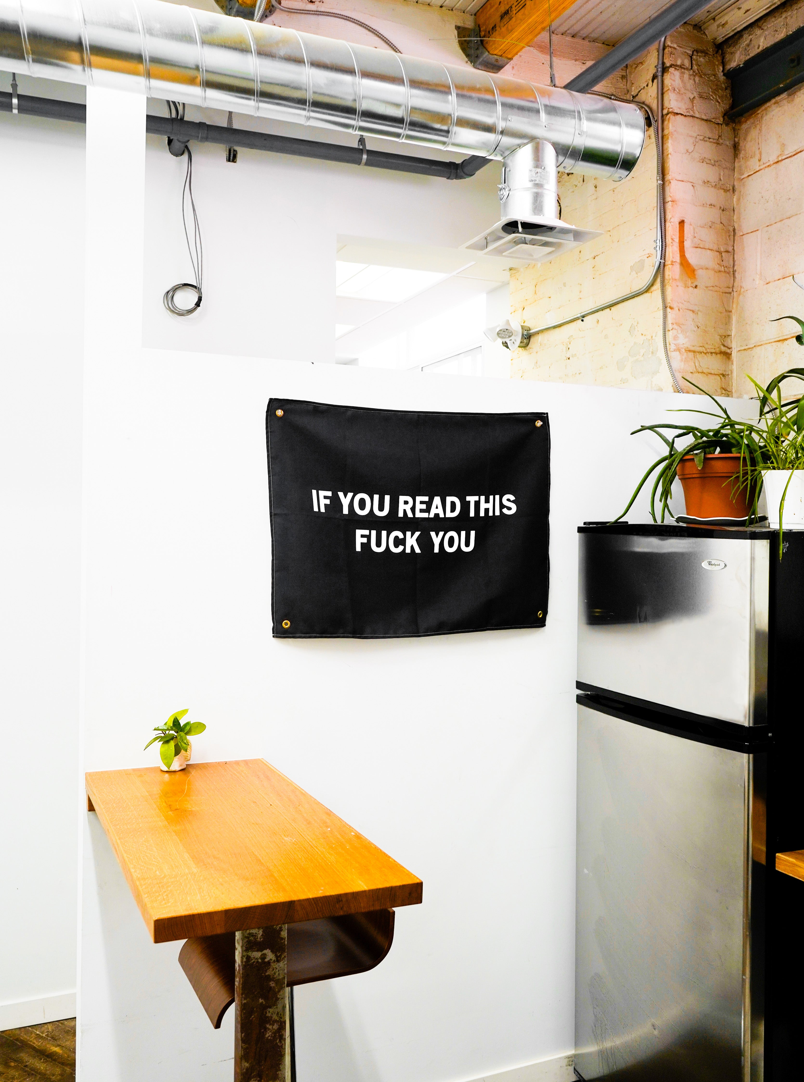 The "Read More" Wall tapestry by No Fun®. Tapestry is black, and features the phrase "IF YOU READ THIS FUCK YOU" in large white letters. There is 1 brass grommet in each corner of the tapestry.  Tapestry is hung on a wall in a kitchen.