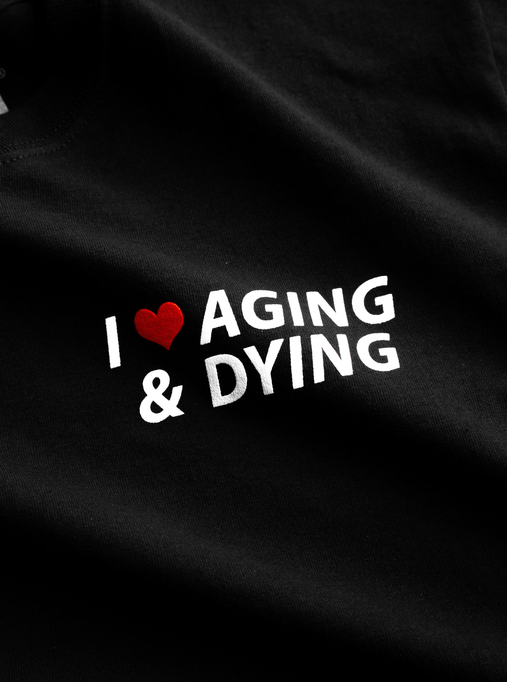 The original "Aging & Dying" T-shirt by No Fun®.  T-shirt is black, and is photographed close up to show the print detail.  The print text reads "I ❤️ Aging & Dying" and is located on the front of the T-shirt.   The text is white, with a red heart.