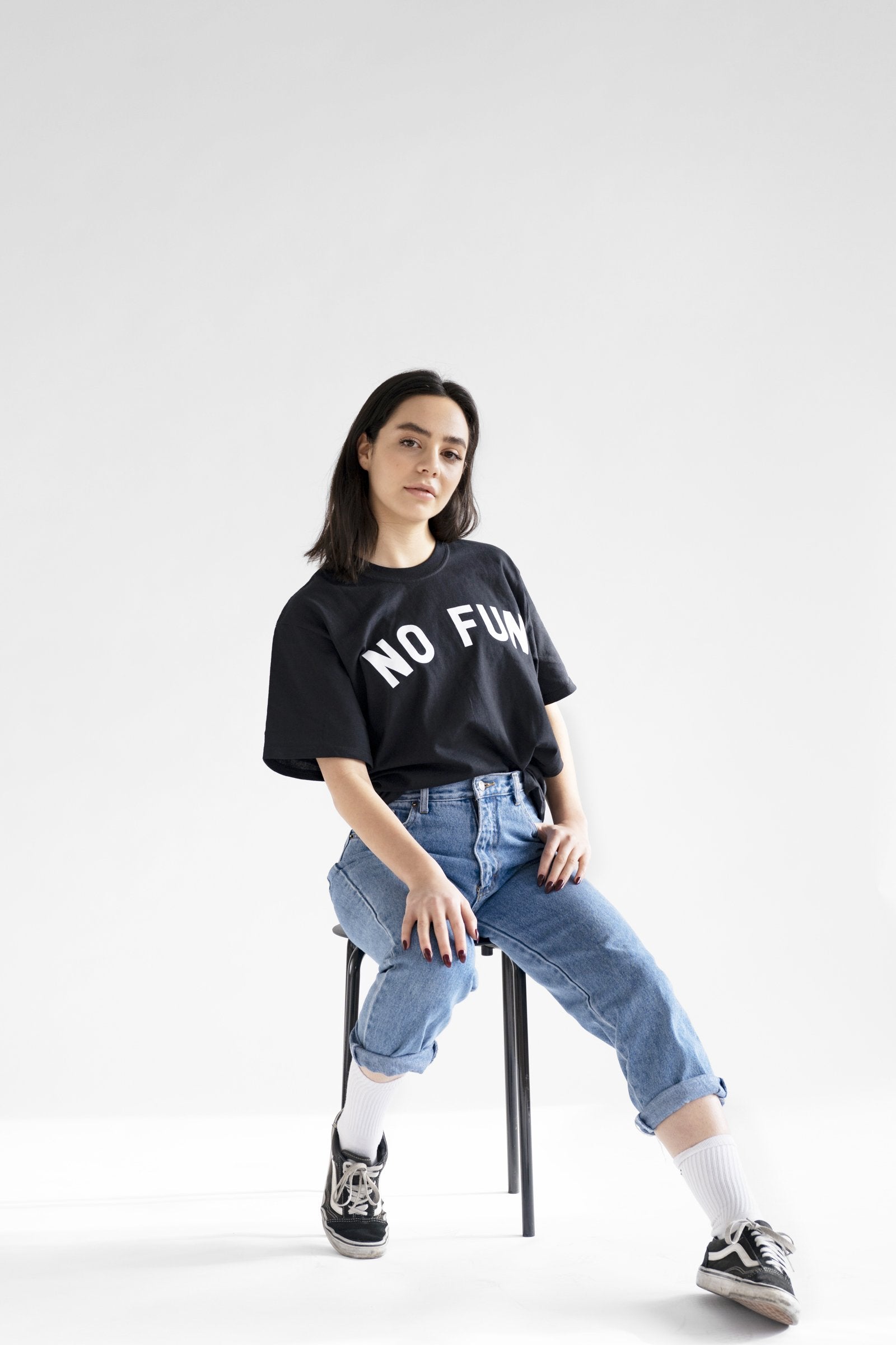 "No Fun®" T-shirt being worn by a female model.  She is also wearing blue jeans, white socks, and black shoes.  She is seated in front of a white background.