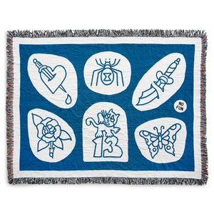 The No Fun® "Flash Sheet" Woven Blanket.  The blanket is blue and white, and photographed against a white background.  The design of the blanket is inspired by traditional tattoo flash sheet designs.  There are six images within the blanket and they include: A heart being pierced by a dagger, a black widow spider, a 2 part dagger, a rose, a cat standing atop the number 13, and a butterfly.  There is also a small "No Fun®" logo in black on the right side.