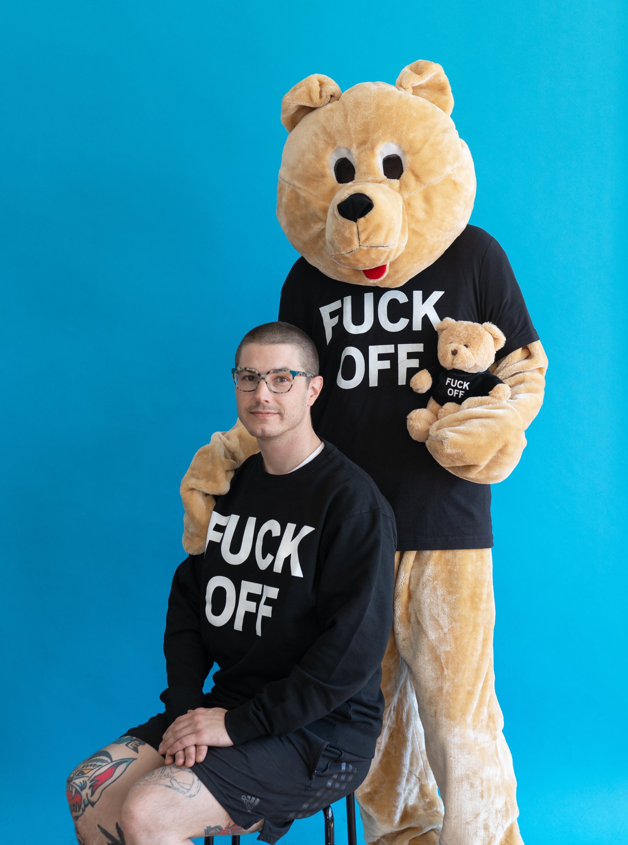 Photo of a model wearing the "Fuck Off" Crewneck by No Fun®.  Model is seated on a stool in front of a blue background.  There is a model behind him wearing a teddy bear costume.