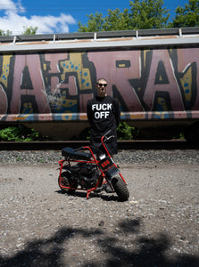 Photo of a model wearing the "Fuck Off" Crewneck by No Fun®.  He is standing behind a red mini bike, and there is a train passing by behind him.