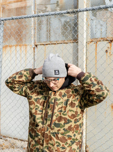 The classic "No Fun" labelled beanie in Heather Grey.  There is a small, black, woven label that reads "No Fun®" on the cuff.  Model is wearing the beanie with a camo jacket.  His arms are up as he is adjusting his hoodie.  He is standing in an industrial area with a chainlink fence behind him