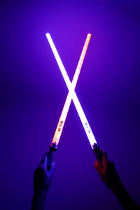 A photo of two hands holding two novelty light up swords.  The light from the swords is illuminating the background in a purple colour.  The sword in the left hand is red , while the right hand is holding blue.  They are crossed in the middle and "NO FUN®" logos are visible to the camera.