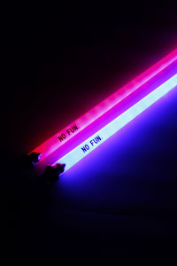 A close up photo of two light up swords showcasing the "NO FUN®" logos close to the handle.  The novelty sword on the left is illuminating the dark background with red light, while the novelty sword on the right is illuminating the dark background with blue light.