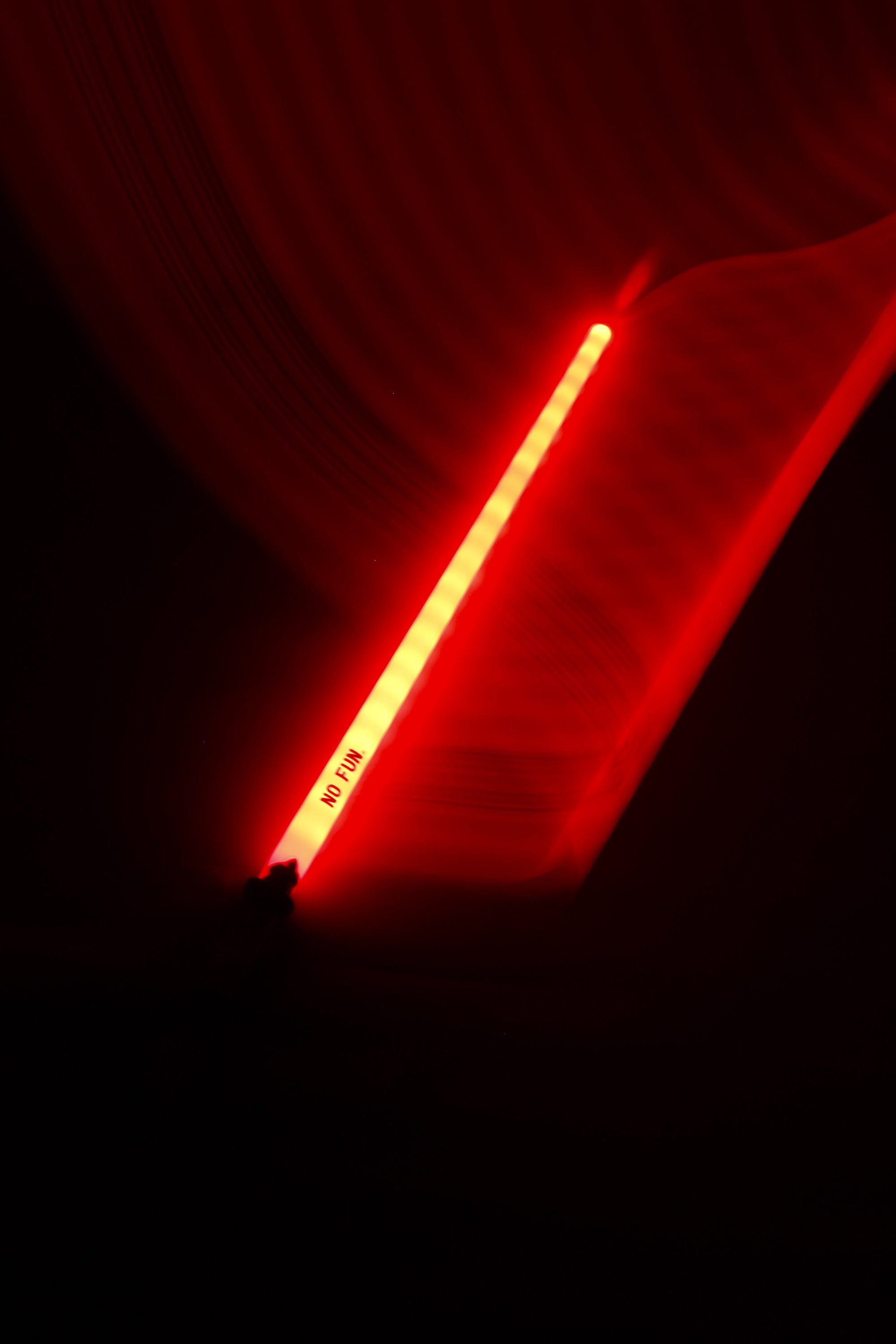 A photo of a red novelty sword illuminating a dark background.  There is a light trail flowing from the sword to the top of the image, showcasing the colour.  The "NO FUN®" logo near the handle is visible to the camera.