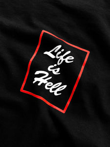 Close up detail of the graphic on the "Life is Hell" T shirt.  The white text in the design reads "life is Hell" in a brush script font, and has a red stroke rectangle around it.  T shirt is slightly wrinkled to show shadows.