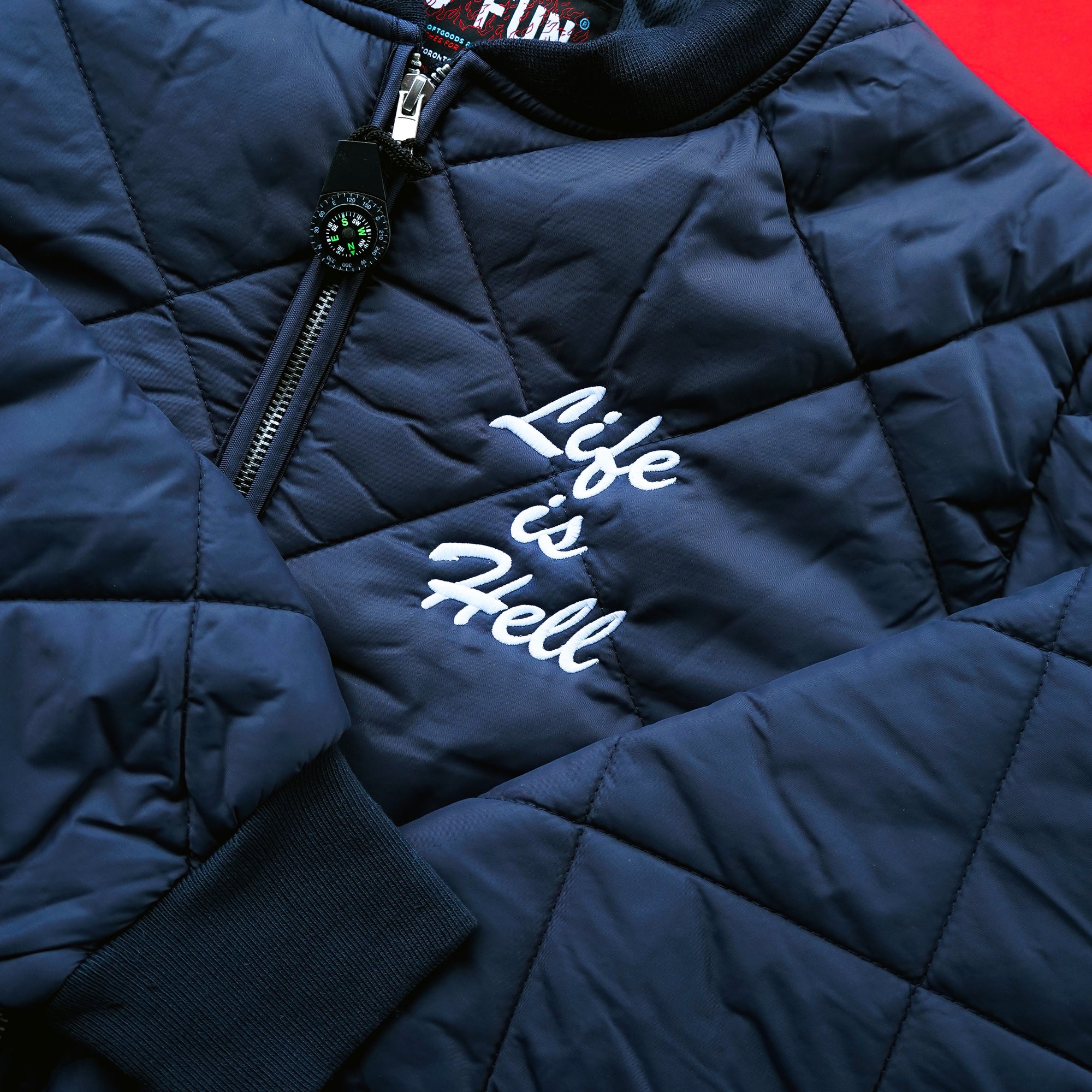 Product photo of the official No Fun® "Life Is Hell" Quilted Bomber Jacket in the navy colourway. The product is photographed on a red background, with the sleeves of the jacket folded over. The jacket features the text "Life Is Hell" embroidered on the front of the jacket in white. The jacket also features a zipper on the front, and full quilting on the entire outer shell.
