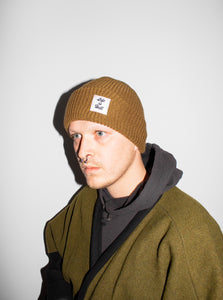 Photo of the "Life is Hell" Beanie by No Fun®.  Product is photographed on a model against a white background.  Model is wearing an olive coloured jacket, grey hoodie, and the Desert Tan colorway of the Beanie.  The beanie features a small white woven label on the cuff that reads "Life Is Hell".  Label is white, with black text, and red contrasting stitching.