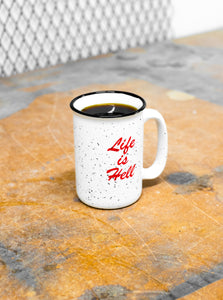 Photo of the "Life Is Hell" Ceramic Camp mug on a scratched, wooden, work table.  There is some metal grating in the back of the image.  The mug is in the center of the image and is filled with black coffee.  The red, "Life Is Hell" print is facing the camera.