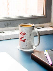 The "Life Is Hell" Ceramic Camp mug on a blue desk.  There is a window behind the mug, and a laptop can be seen in the bottom left corner of the image.  There is also a sketch book and pen in the foreground.  The mug is on a clear coaster, and the "Life Is Hell" graphic in red is visible.