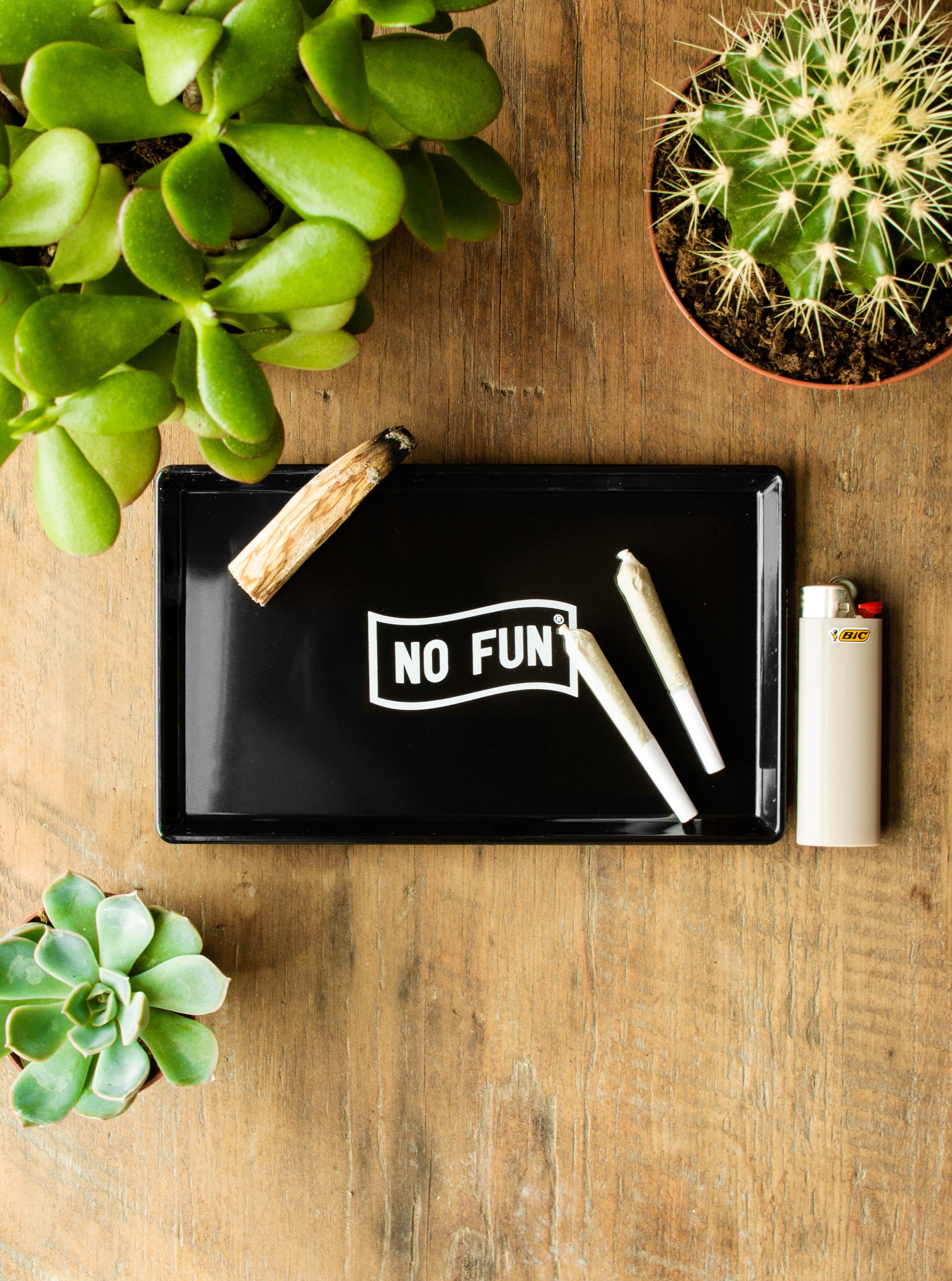 The No Fun® Multi-Purpose Tray on a wooden table.  The tray is surrounded by various succulents.  There is a grey lighter on the right side of the tray.  There are 2 joints, and a piece of Palo Santo laying in the tray.