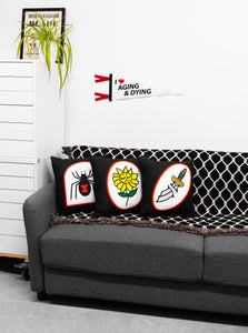 Varios pillows by No Fun® on a couch in a white room.  The couch is grey, and with a white shelf on the left.  There is a plant and print on top of the shelf.  An "Aging & Dying" pennant is hanging on the wall above the couch.  There is a 'Chainlink" woven blanket draped over the back of the couch.