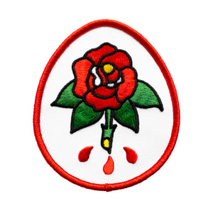 The "Rose" Patch by No Fun®.  Patch is white, with a red border and is an oval shape.  There is a cartoon rose found in the center of the patch.  There are 3 red drips emanating from the roses stem.