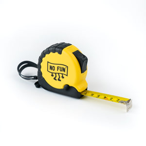 The "WORK" Tape Measure by No Fun®. The body of the tape measure is yellow, with black accent trim. The photo shows the product against a white background with the tape measure visible. This side of the product features a "No Fun®" logo in the shape of a banner. The logo also has arms and legs. You can also see the brake on the tape measure, as well as the locking mechanism on the top right.