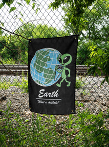 The "Tourist" Wall tapestry by No Fun®. Tapestry is black, and features a cartoon alien with a camera positioned in front of a globe. The phrase "Earth 'What a shithole!'" is underneath the globe. There is 1 brass grommet in each corner of the tapestry.  Tapestry is hanging on a chainlink fence surrounded by foliage.  There are train tracks in the distance.