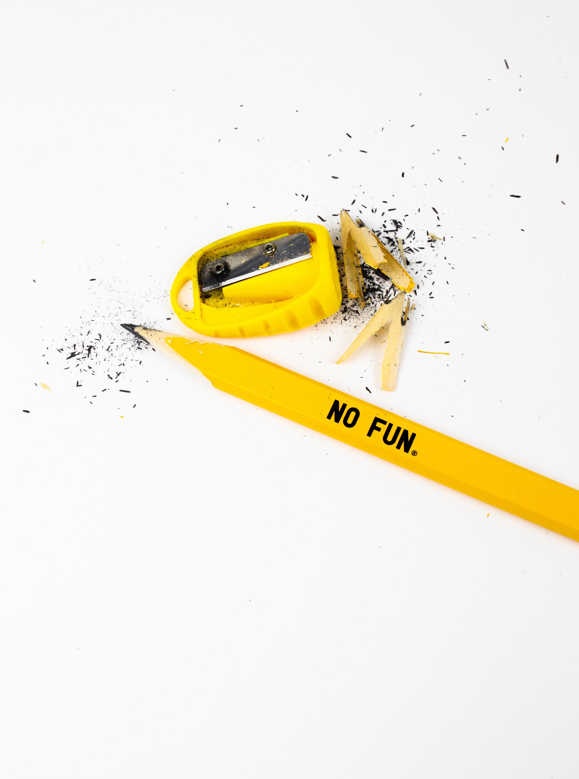 A yellow carpenter pencil and sharpener.  Graphite shards and pencil shavings are found near the sharpener.  There is a small No Fun® logo in black, on the center of the pencil.